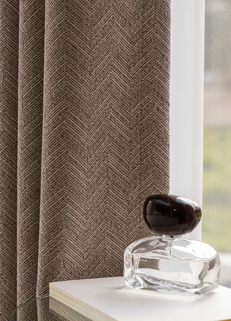 a clear glass perfume bottle with a dark cap, placed on a white surface next to a window with a brown herringbone-patterned curtain.
