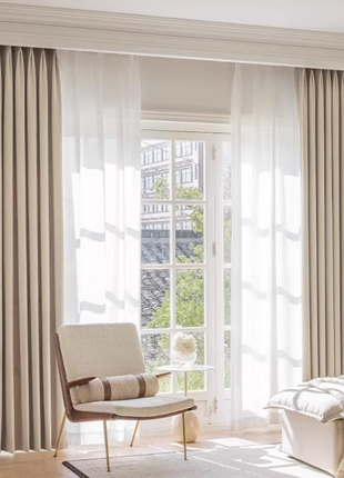 a well-lit living room with a comfortable chair, a glass table, and large windows with sheer curtains.