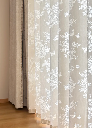a set of white curtains with a floral pattern, hanging on a wooden floor. The curtains are partially open, allowing natural light to enter the room.