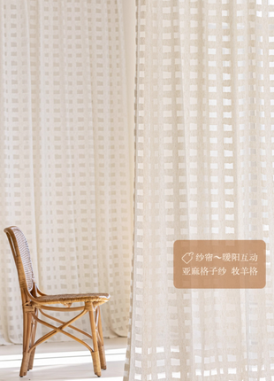 A wooden chair with a woven seat and backrest is positioned in front of a white curtain with a pattern of small squares.