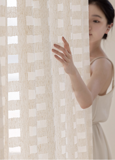 A woman in a white dress stands behind a beige curtain, her hand reaching out to the curtain.