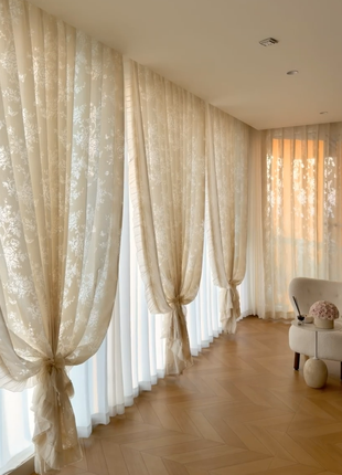 a spacious room with a long wooden floor and three beige curtains hanging from the ceiling, creating a sense of depth and dimension. A white armchair is placed in the corner of the room, and a vase of flowers is visible on the floor.