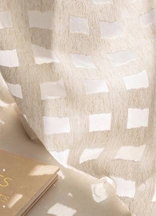 a close-up view of a beige fabric with a white pattern, partially draped over a surface. There is a small book with gold lettering on the left side of the frame.