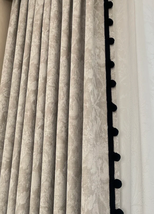 a close-up view of a set of curtains with a gray and white floral pattern, featuring a black trim with pom-poms.