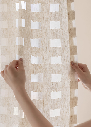A person's hands are holding a beige curtain with a pattern of white squares.