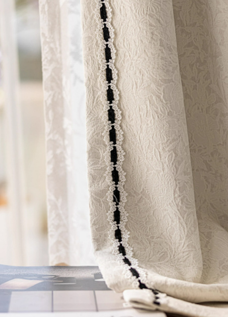 a close-up view of a white curtain with a black lace trim, hanging in front of a window. The curtain appears to be made of a delicate fabric, possibly lace, and is adorned with a decorative black lace trim. The curtain is partially drawn, revealing a glimpse of the window behind it. The background is blurred, drawing attention to the curtain and its details.