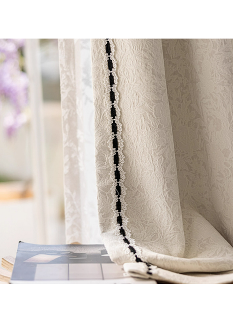 a close-up view of a white curtain with a black lace trim, hanging in front of a window. The curtain appears to be made of a delicate fabric, possibly lace, and is adorned with a decorative black lace trim. The curtain is partially drawn, revealing a glimpse of the window behind it. The background is blurred, drawing attention to the curtain and its details.