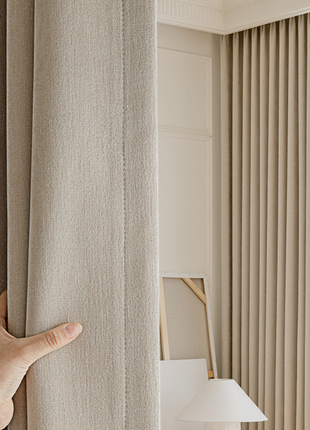 A hand is holding a beige curtain, partially covering a window with vertical blinds.