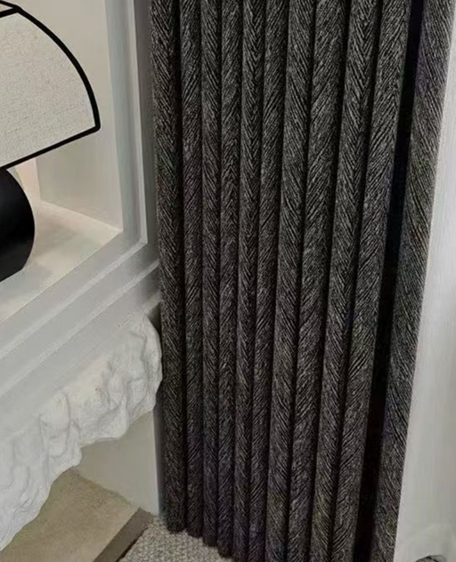 a close-up view of a dark grey curtain with a herringbone pattern, hanging next to a white wall.