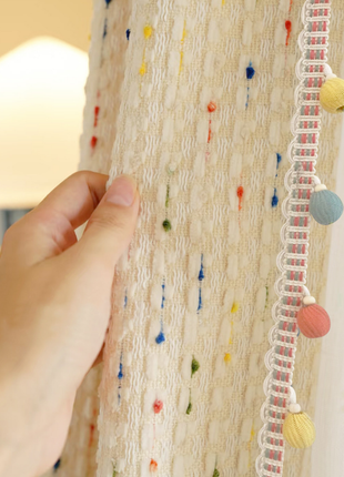 A hand is holding a colorful, textured fabric with multicolored balls hanging from it.