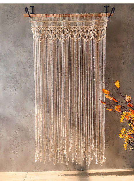 Rideau macramé bohème écru tissé main sur tringle en fer forgé devant un mur beige